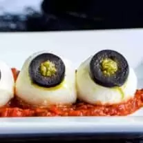 Three mozzarella balls with black olive eyes, resembling Halloween eyeballs, on a white plate.