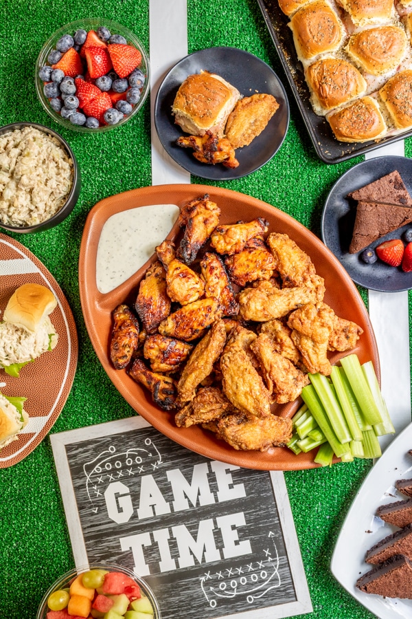 Top down view of football party food table with chicken wings in the center, fresh fruit, and sliders. 