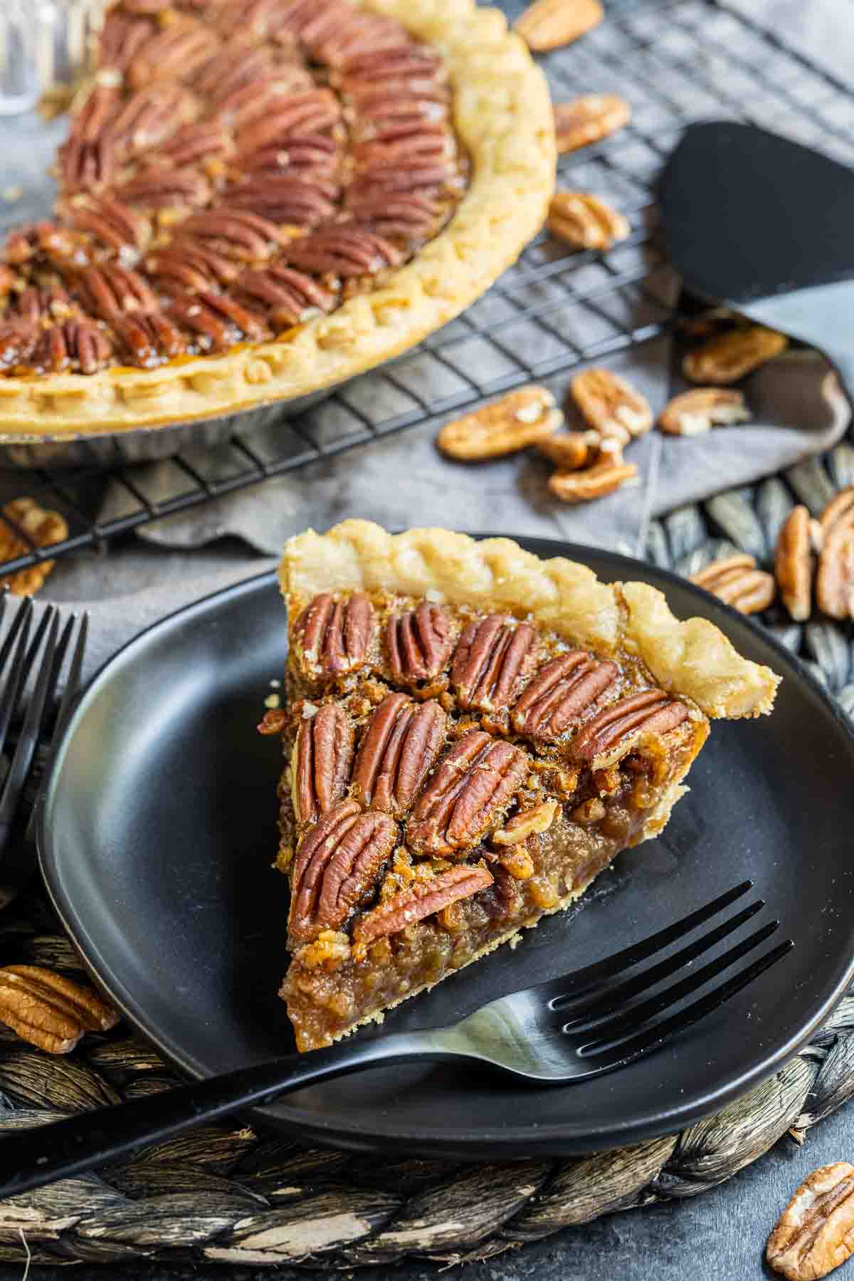 slice of pecan pie on a black plate with a black fork and pie cooling on rack