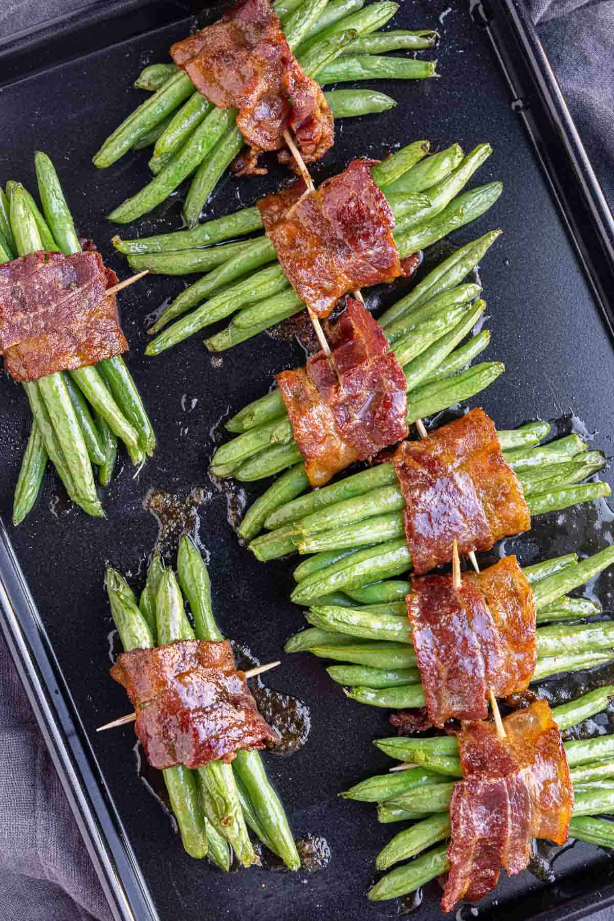 Bacon Wrapped Green Beans bundled together with toothpicks on a black sheetpan