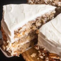 A slice of carrot cake with cream cheese frosting and pecan nuts on a wooden surface.