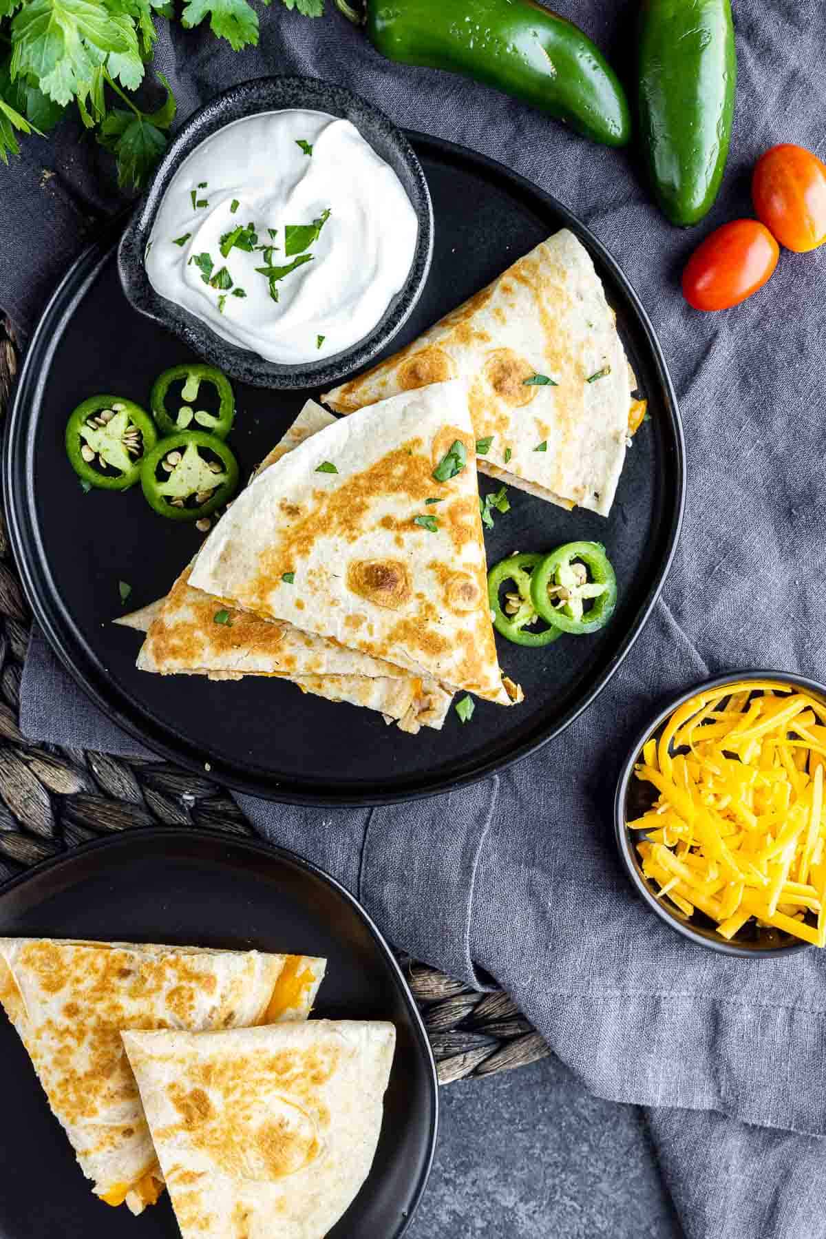 two black plates with Chicken Quesadillas, small bowl of sour cream, jalapeno slices and shredded cheese