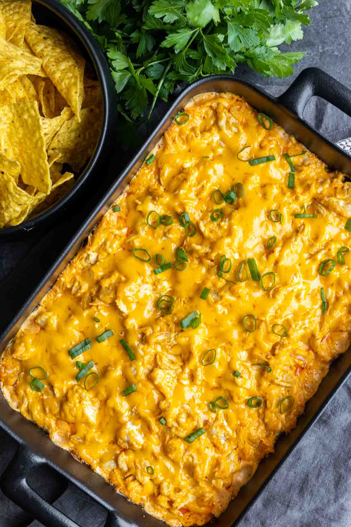Oven Buffalo Chicken Dip in a black casserole dish with parsley and a bowl of chips