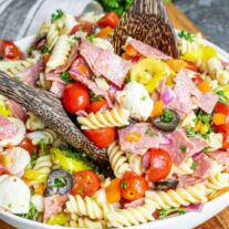 A vibrant pasta salad with tomatoes, olives, peppers, salami, and mozzarella, mixed with a wooden serving utensil.