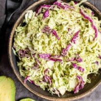 A bowl of coleslaw with red cabbage and green cabbage served with lime and avocado on the side.