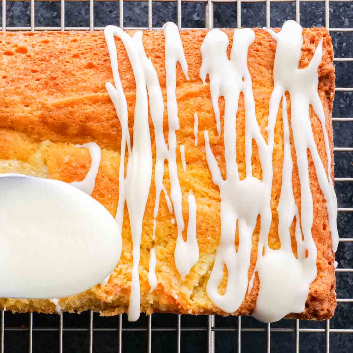 A lemon loaf cake with icing and a spoon on a cooling rack.