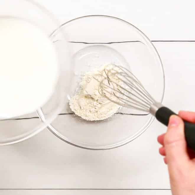 A person whisking flour and milk in a glass bowl to make Creamed Corn