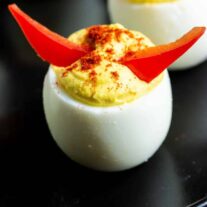 Deviled eggs topped with red and yellow peppers on a black plate.
