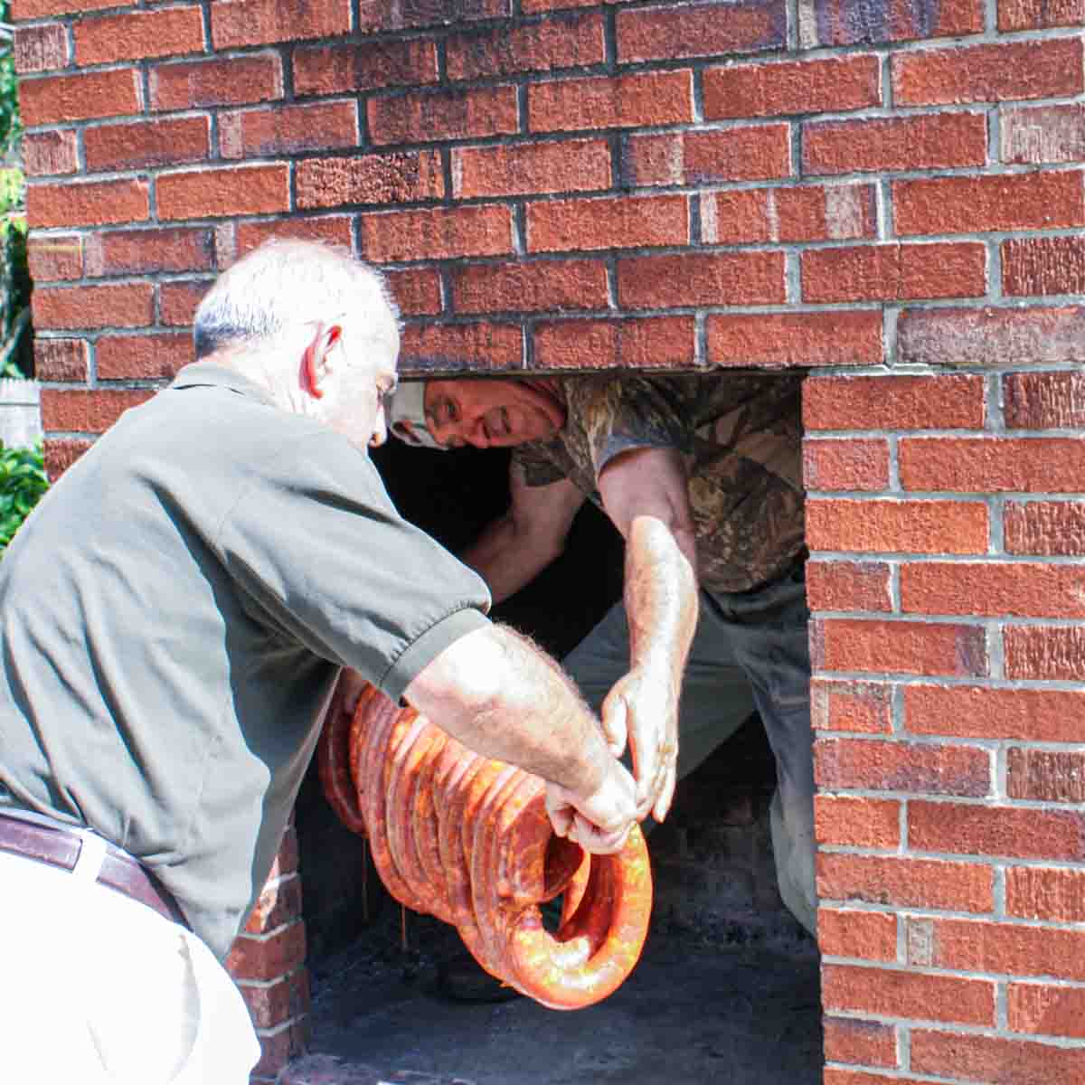 Two men putting Portuguese sausage into a brick fireplace.