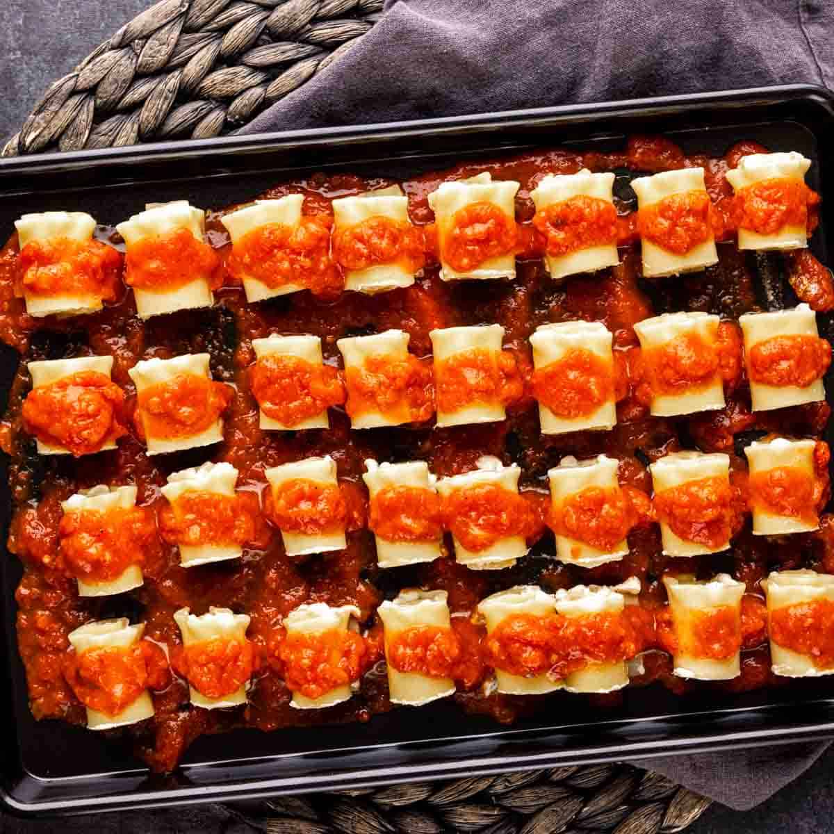 A black baking sheet with a tray of mini lasagna appetizer with sauce on it.