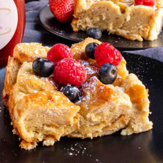 A slice of Overnight French Toast Bake with berries and syrup on a black plate.