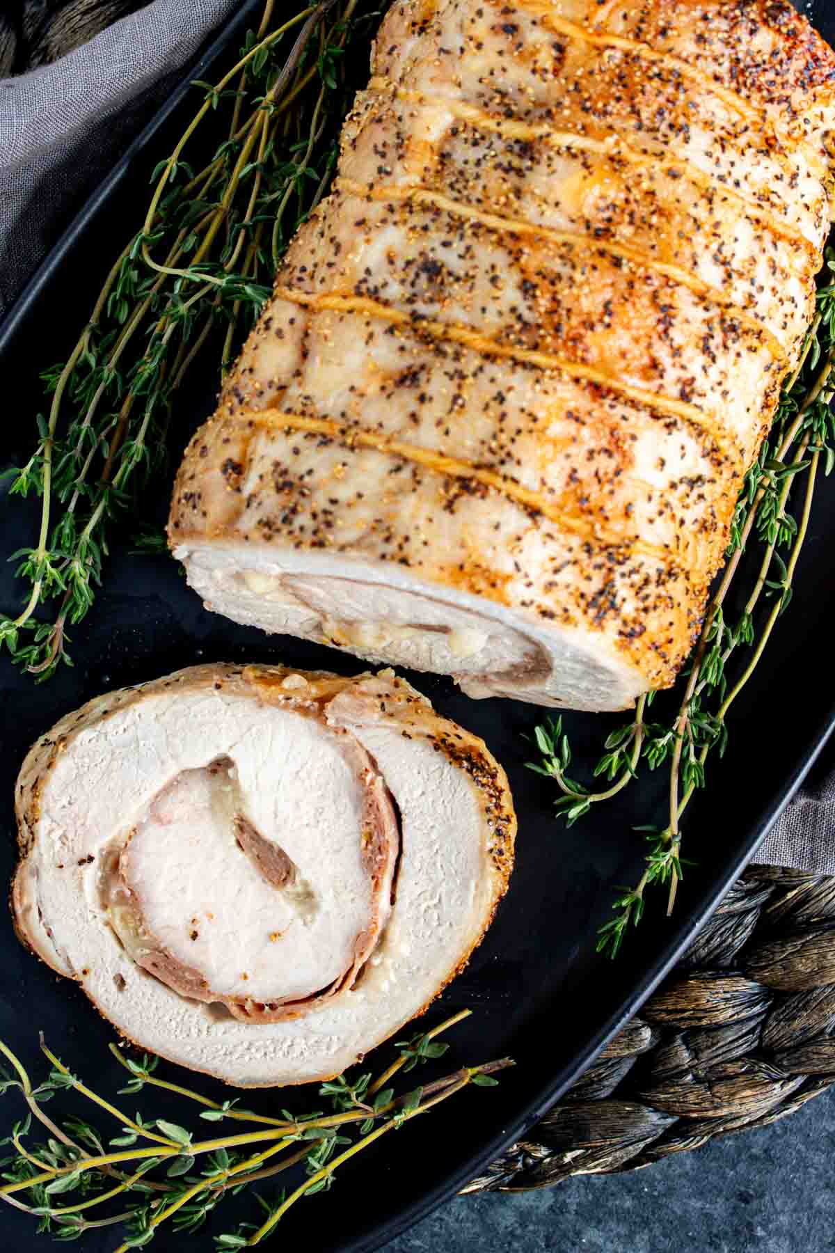 A Provolone and Prosciutto Stuffed Pork Loin on a black plate with thyme sprigs.