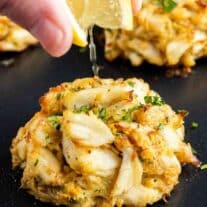 A person drizzling lemon juice on a crab cake.