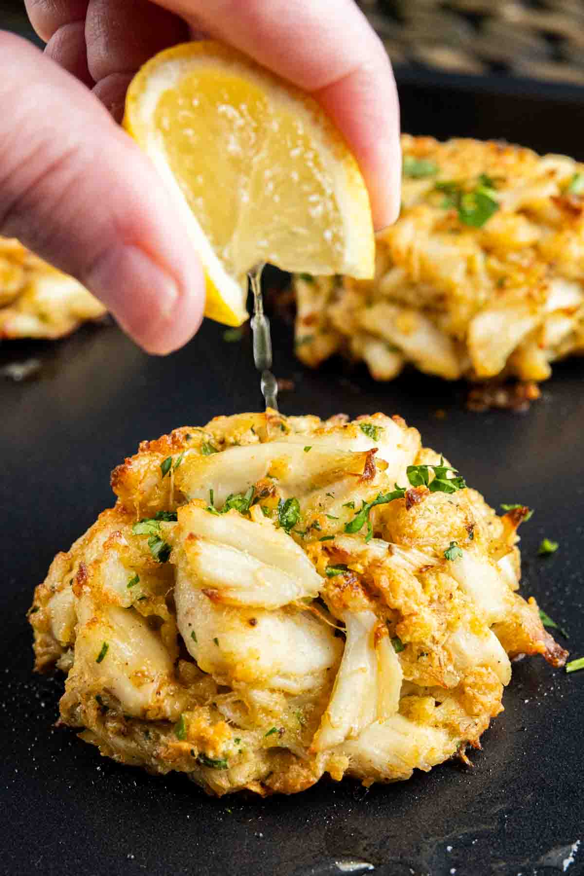 A person is drizzling lemon juice on some Maryland Crab Cakes.