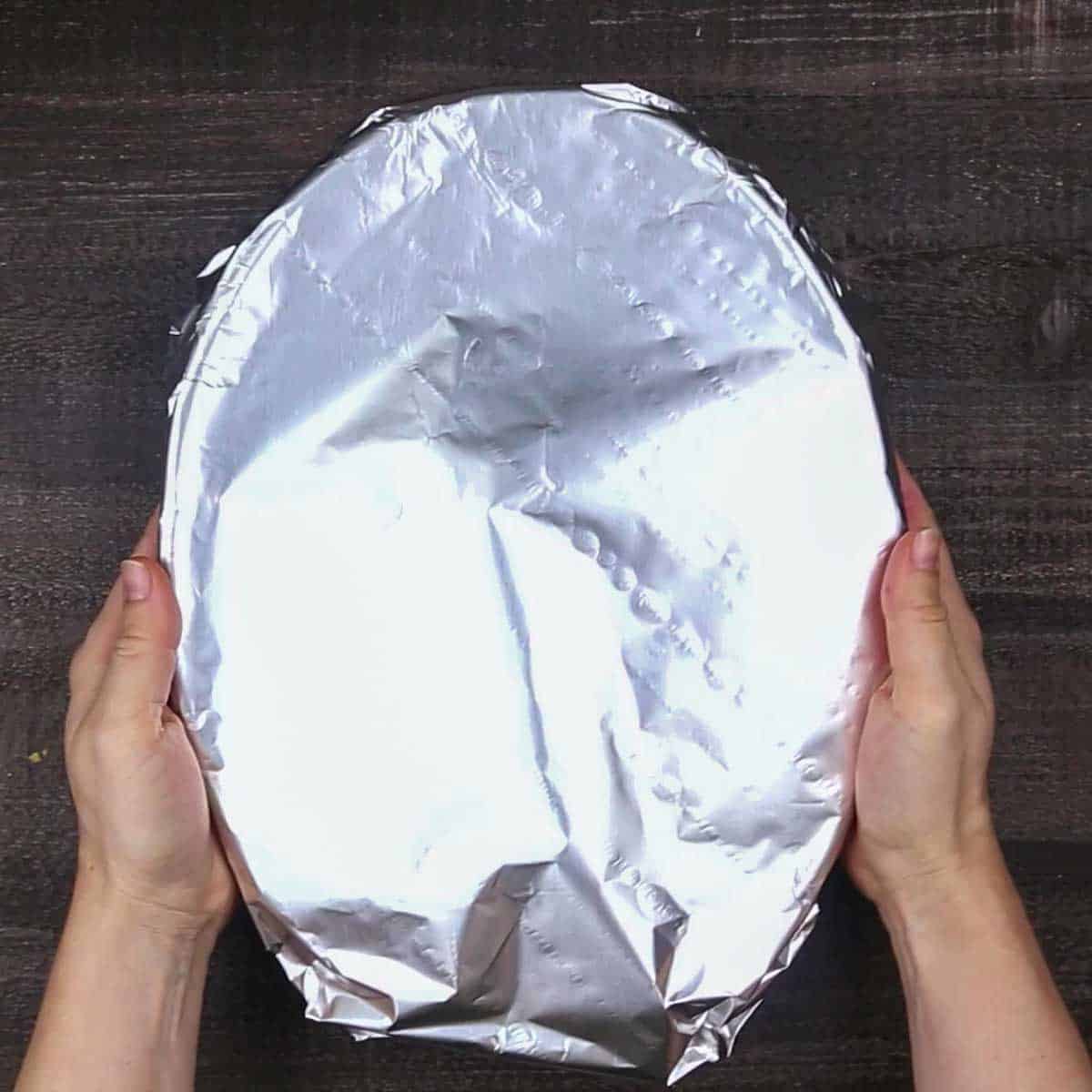A person holding a piece of foil on a wooden table covering Beef Enchilada Casserole