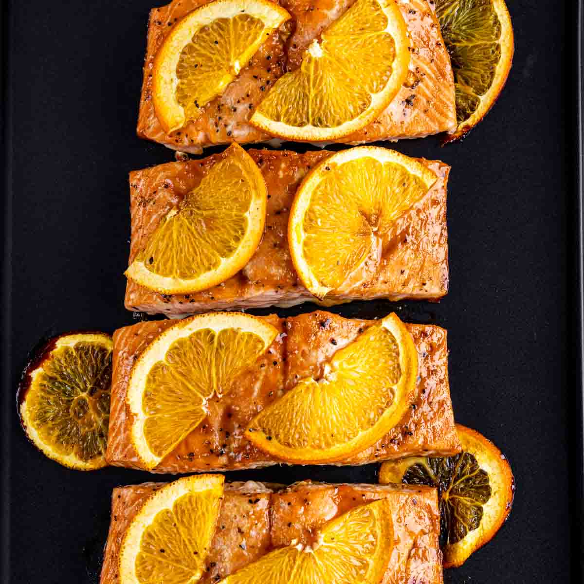 Four pieces of Orange Glazed Salmon topped with orange slices on a black surface.