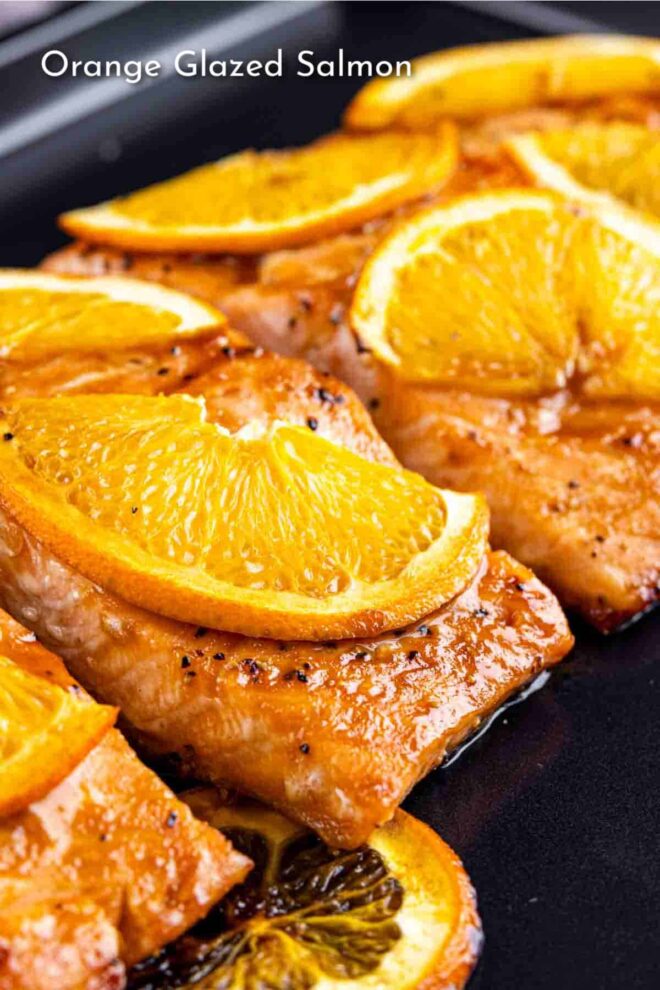 Slices of orange glazed salmon garnished with orange slices on a black cooking surface.