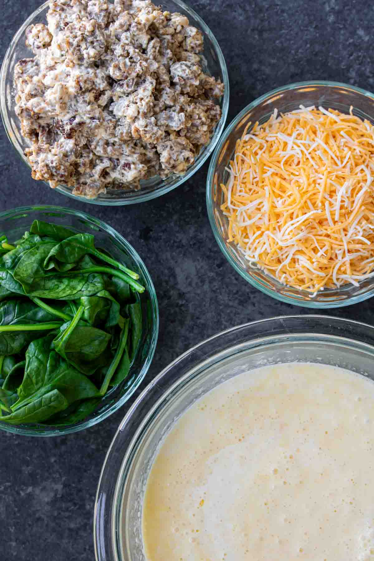 Ingredients for a recipe arranged on a countertop, including a bowl of seasoned ground meat, shredded cheese, fresh spinach, and a eggs for Spinach and Sausage Quiche
