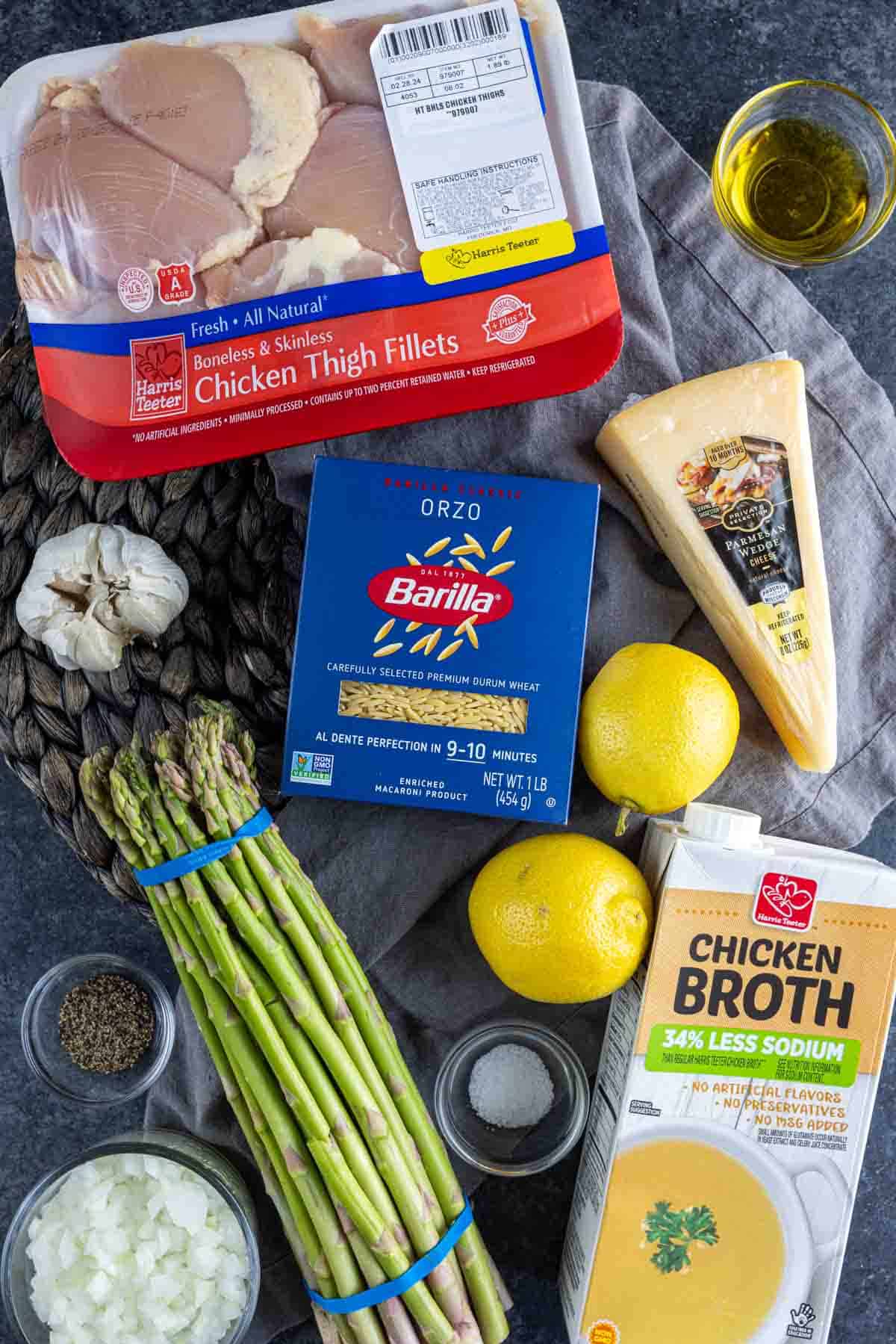 Parmesan Chicken Orzo Ingredients for a recipe including chicken thigh fillets, orzo pasta, parmesan cheese, lemons, asparagus, garlic, chicken broth, olive oil, and spices on a dark surface.