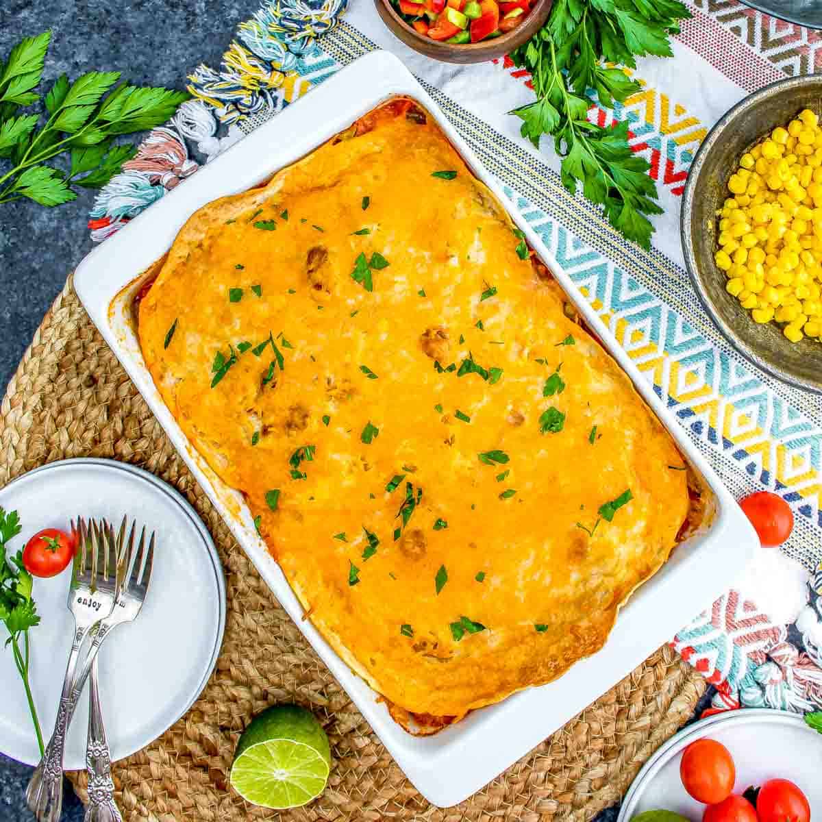 Overhead view of a homemade Chicken Tortilla Casserole in a white dish surrounded by fresh herbs, a bowl of corn, and salad, served on a colorful tablecloth.