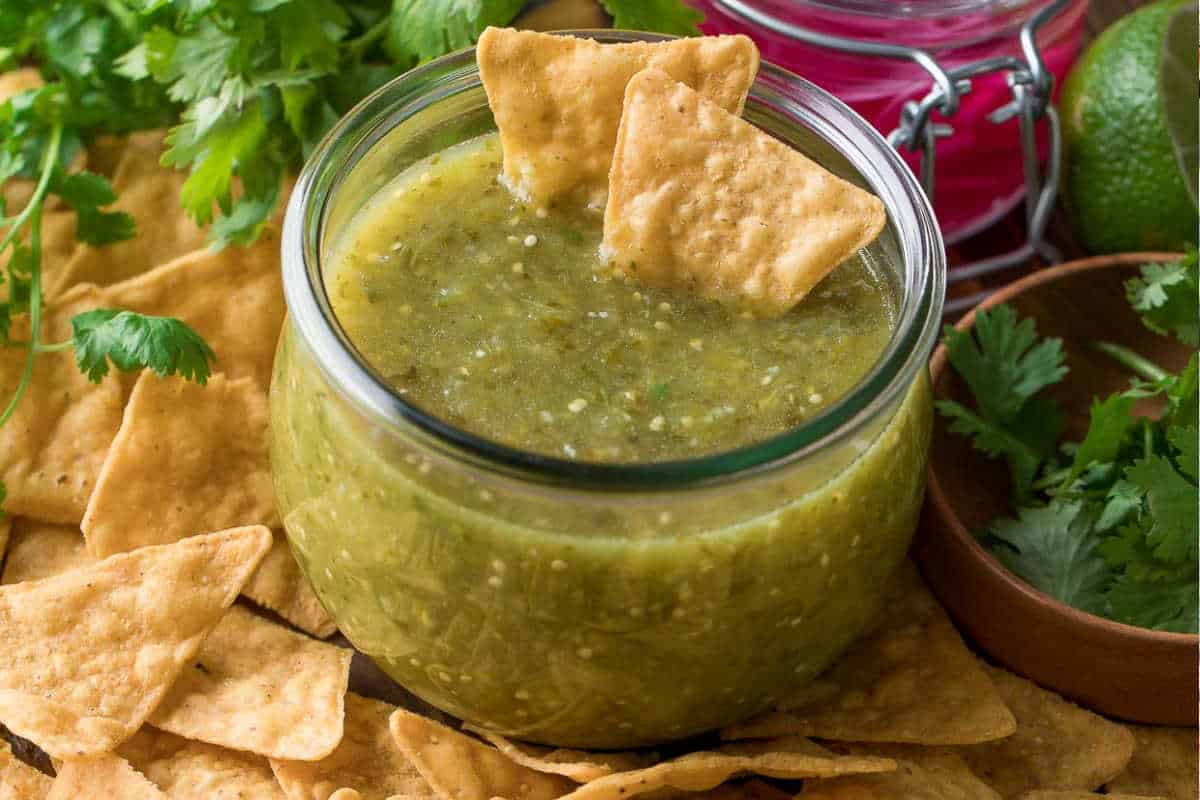 A glass jar filled with salsa verde, surrounded by tortilla chips and fresh cilantro, on a wooden surface.