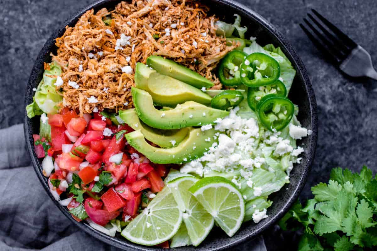 A colorful taco salad with shredded chicken, avocado slices, tomatoes, jalapeños, crumbled cheese, and lime wedges in a black bowl.