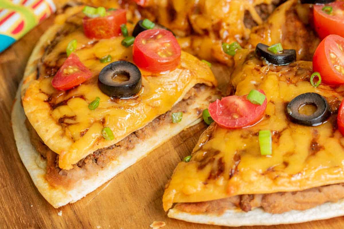 Sliced cheeseburger pizza topped with halved cherry tomatoes, sliced black olives, and chopped green onions on a wooden board.