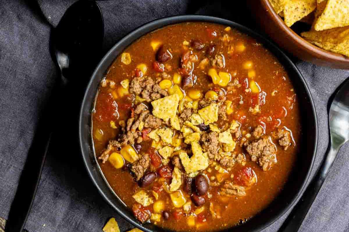 A bowl of taco soup with ground meat, beans, corn, and crushed tortilla chips, served on a dark cloth background.