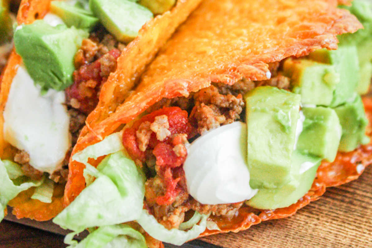 A close-up of a taco with crispy shell filled with ground beef, lettuce, sour cream, and diced avocado on a wooden surface.