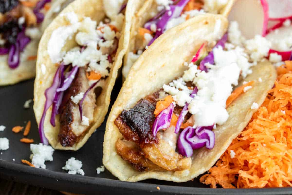 Three pork tacos with purple cabbage, carrots, and crumbled cheese in soft shells, served on a black plate.