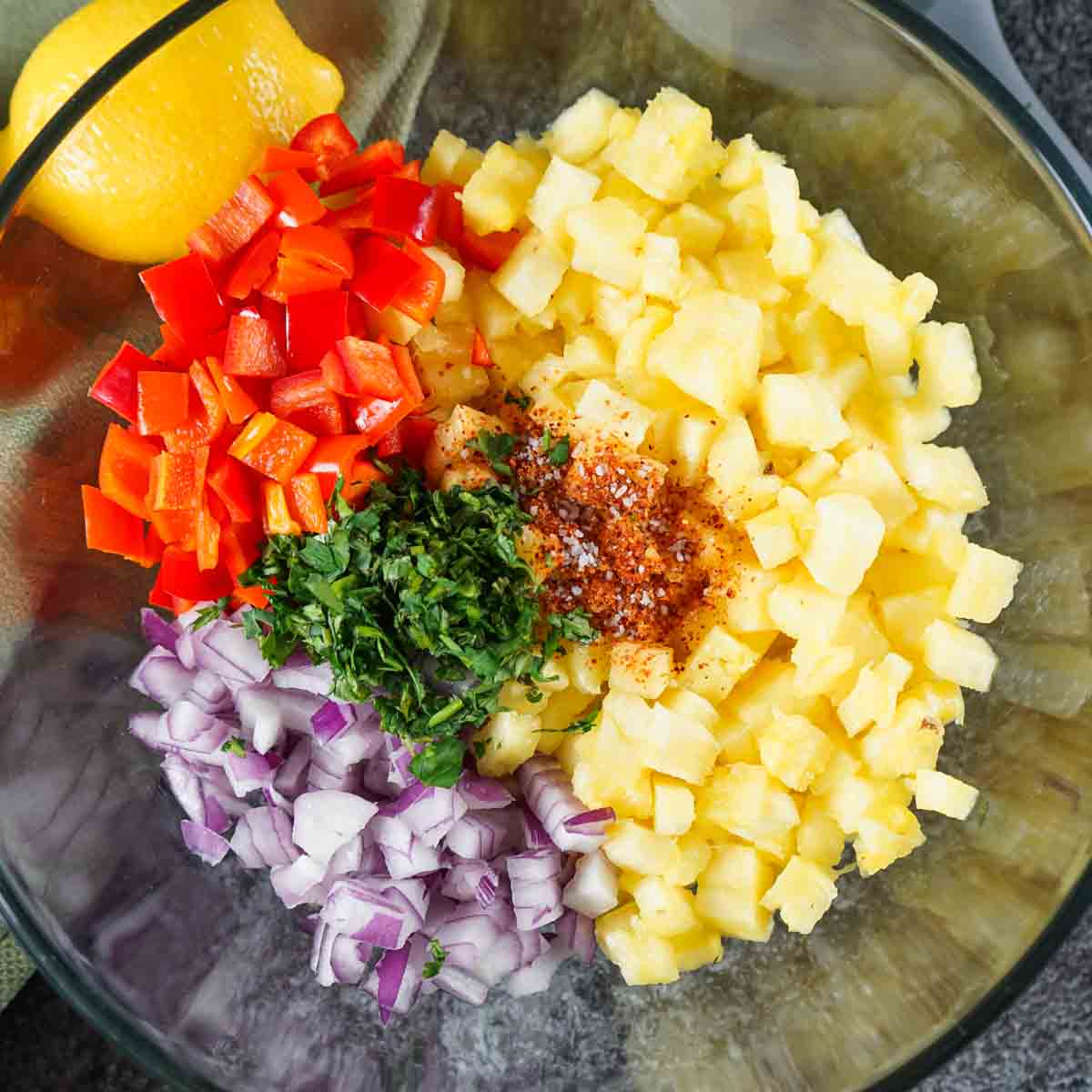A bowl with chopped pineapple, red bell peppers, red onions, cilantro, and spices, with a lemon on the side.
