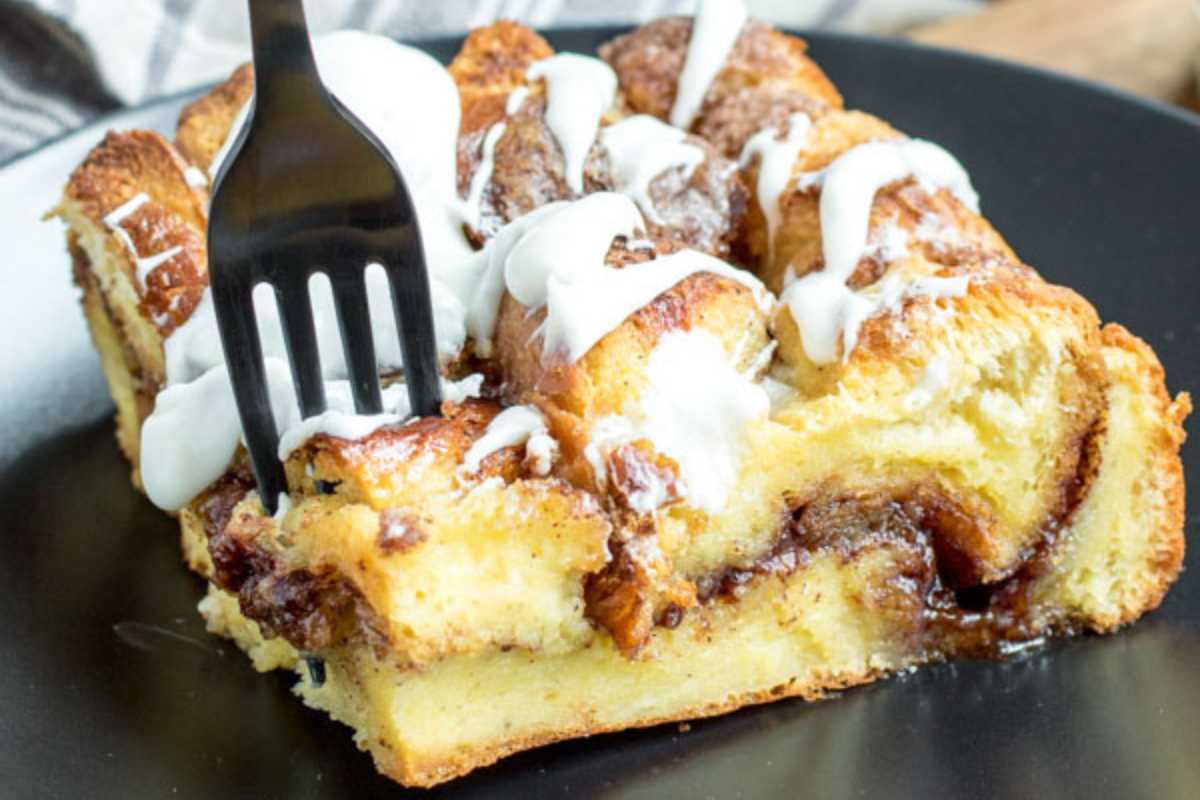 A slice of cinnamon roll french toast bake topped with icing on a black plate with a fork.