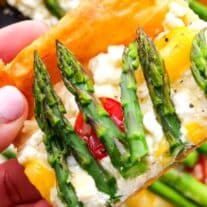 A close-up of a hand holding a slice of vegetable-topped flatbread with asparagus, peppers, and cheese.