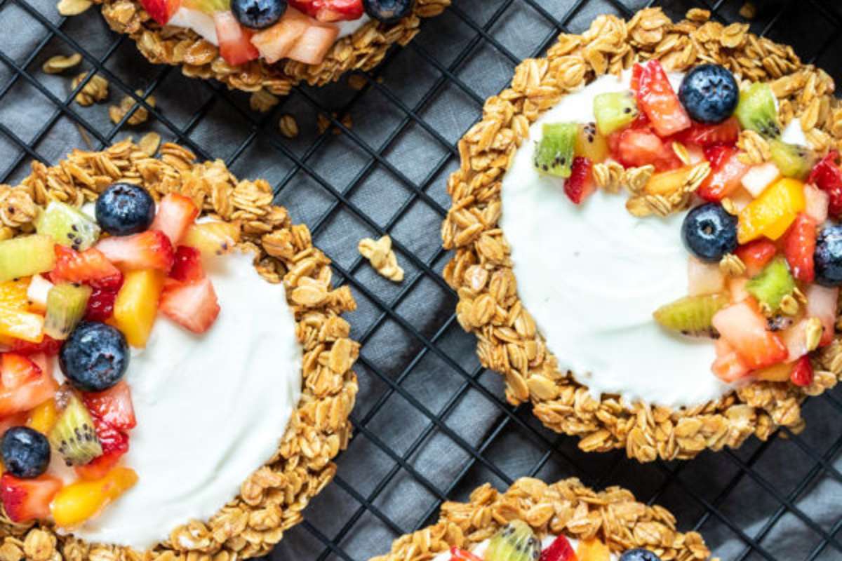 Three granola cups filled with yogurt and topped with assorted fresh fruits, on a dark grid background.