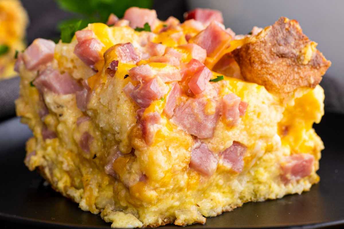 A slice of ham egg and cheese bake topped with diced ham and melted cheese, perfect for celebrating Mother's Day, served with a piece of toast.