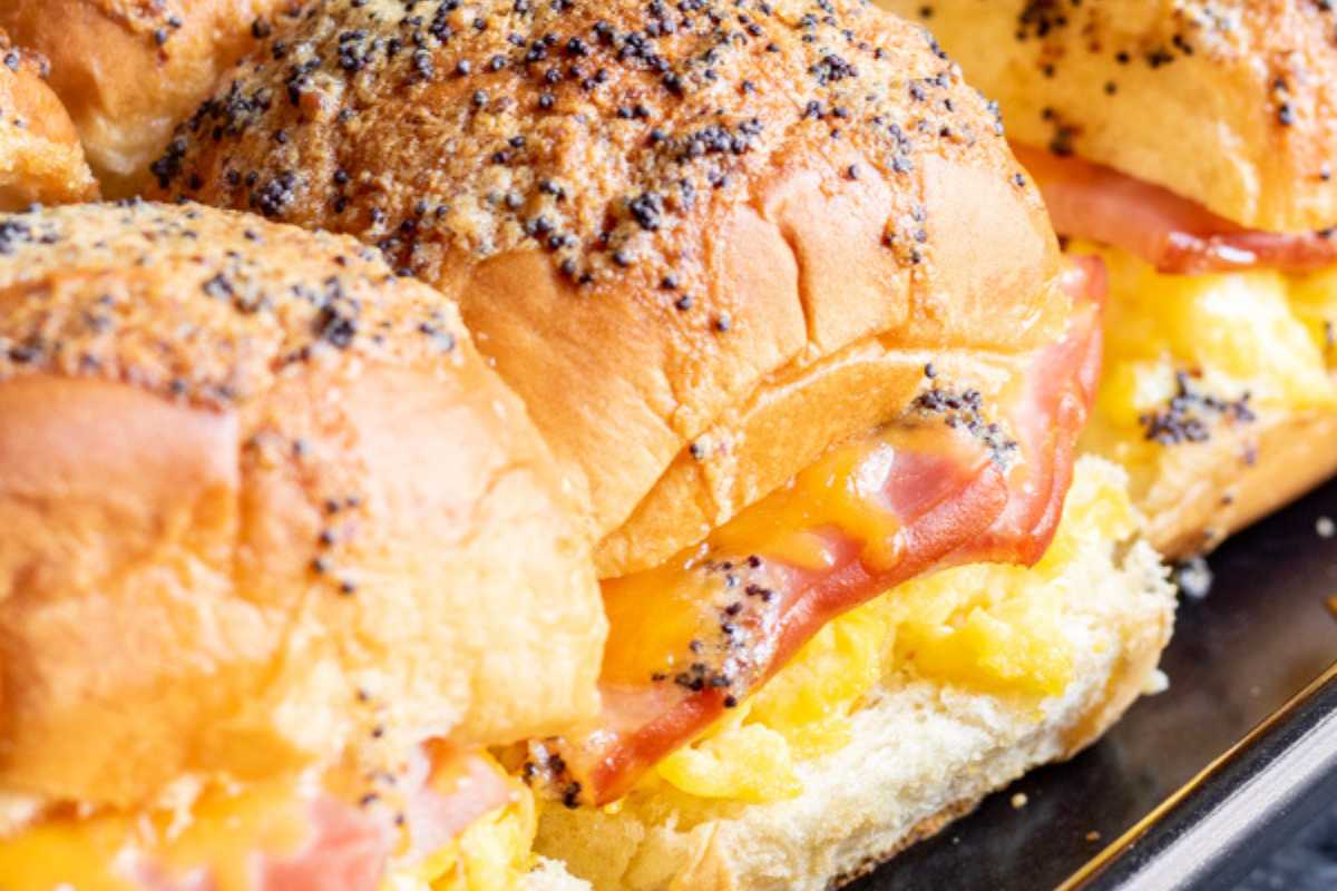 Close-up of a tray with a sandwich made of a poppy seed bun filled with scrambled eggs, melted cheese, and slices of ham.