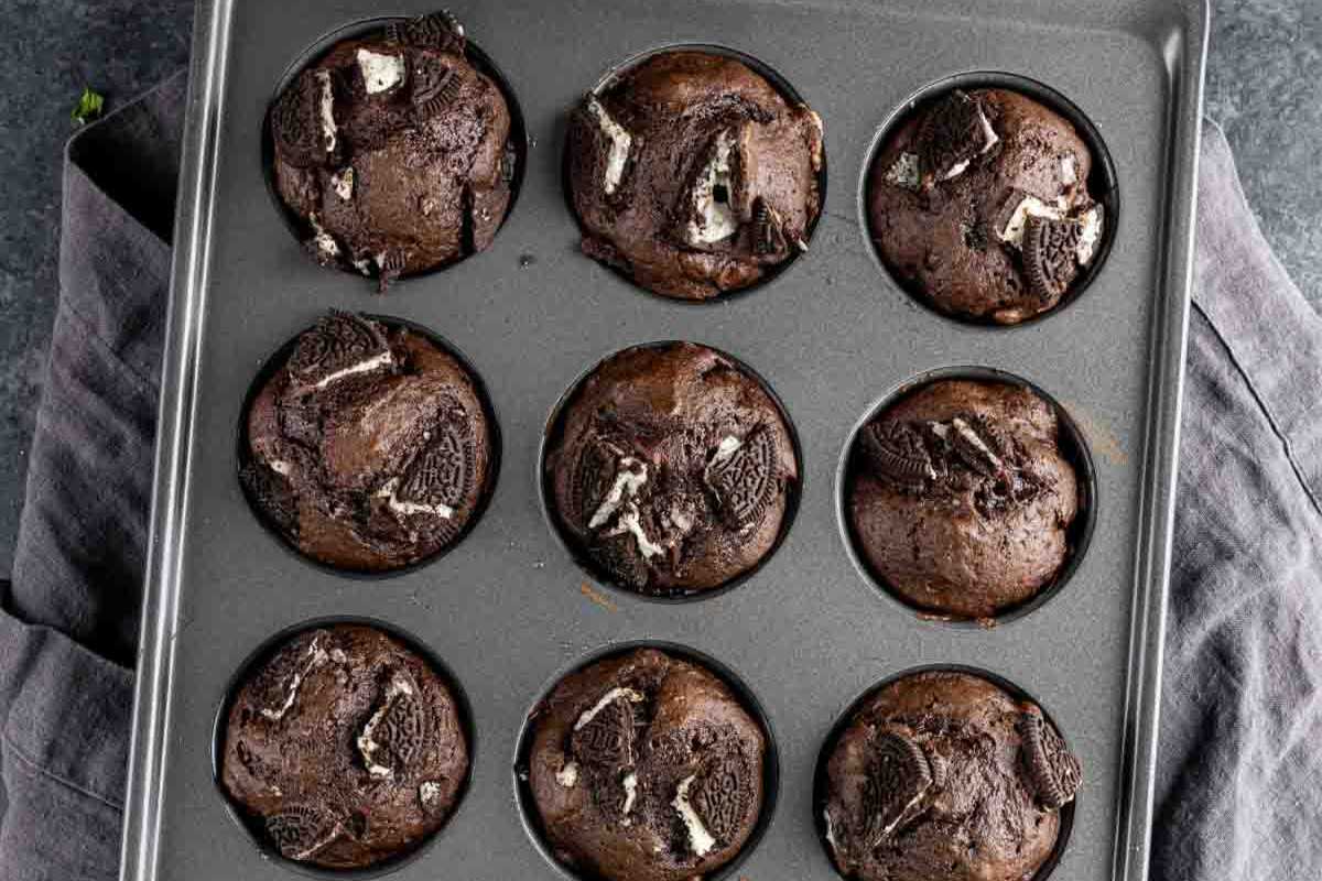 A tray of freshly baked chocolate muffins with chunks of Oreo on top, displayed in a gray muffin tin on a dark countertop.