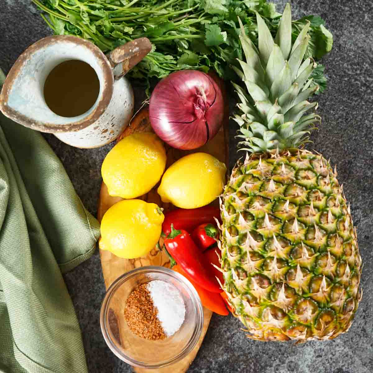 Assortment of fresh ingredients including pineapple, lemons, red peppers, onion, herbs, and seasonings on a gray countertop.