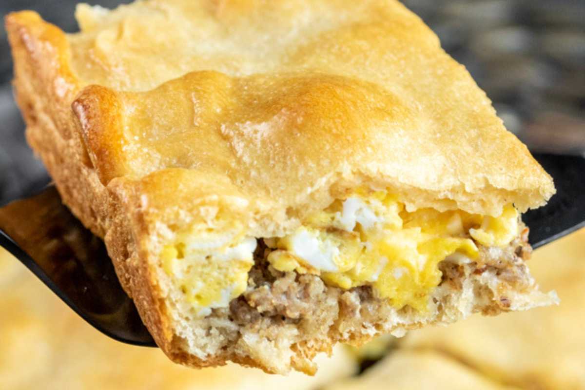 Slice of breakfast pie with layers of egg, sausage, and cheese on a spatula, against a blurred background.
