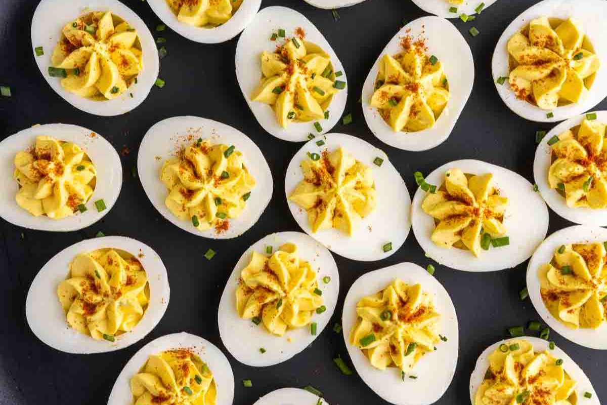 A platter of deviled eggs garnished with paprika and chopped chives on a dark background.
