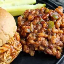 Close-up of a plate with a pulled chicken sandwich on a bun, a side of Baked Beans Recipe with diced green peppers, and pickle spears in the background.