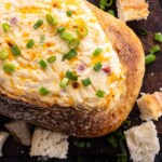 A loaf of bread hollowed and filled with creamy cheese dip, topped with chopped green onions, surrounded by bread pieces on a wooden surface.