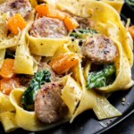Close-up of a plate with pappardelle pasta, sliced sausage, spinach, and chunks of orange squash, garnished with grated cheese and black pepper.