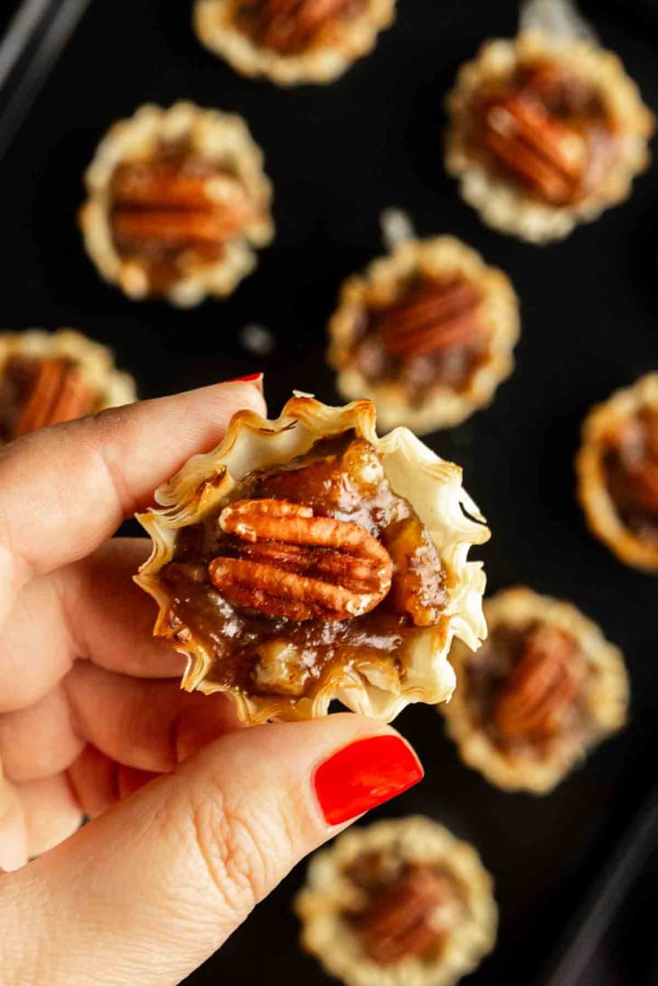 A hand with red nail polish holds a mini pecan pie, featuring a flaky crust and pecan topping; more bite‑sized pecan pie treats are visible on a dark surface in the background.