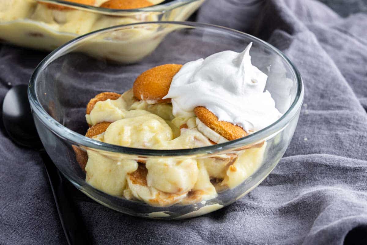 A glass bowl filled with banana pudding, vanilla wafers, and a dollop of whipped cream on top sits on a gray cloth—one of the best easy Thanksgiving desserts for a stress-free holiday treat.