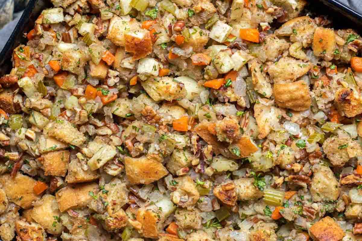 Close-up of a baked bread stuffing dish with visible chunks of bread, diced carrots, celery, onions, herbs, and pecans.
