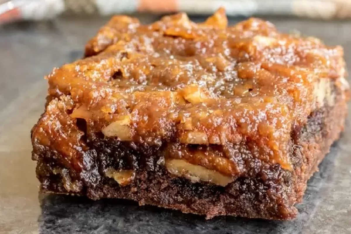 A close-up of a single slice of gooey chocolate and nut dessert bar on a dark surface.