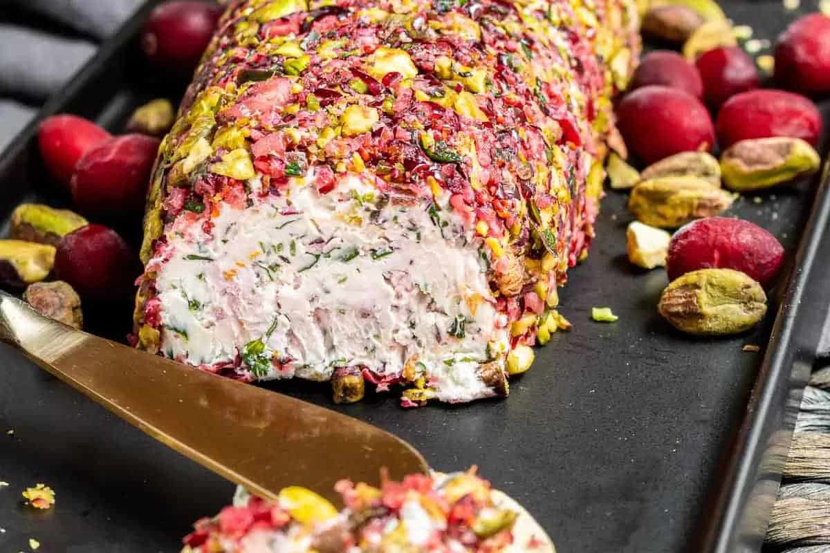 A log-shaped cheese spread coated with chopped pistachios and cranberries, partially sliced, on a black tray with whole pistachios and cranberries.