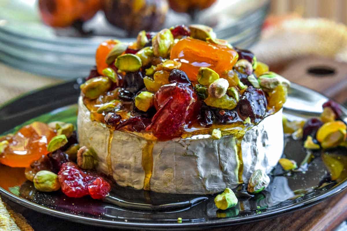 A round of brie cheese topped with dried fruits, pistachios, and honey, served on a black plate with extra toppings around the cheese.