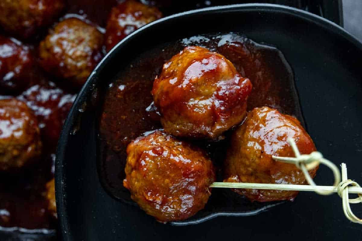 Three glazed meatballs coated in a dark sauce sit on a black plate, with one meatball skewered by a small wooden pick.